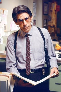 a man wearing glasses and suspenders is reading a book in an office setting,