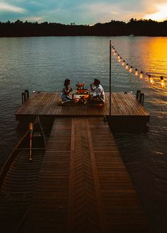 two people sitting on a dock with lights strung across the water and one person eating