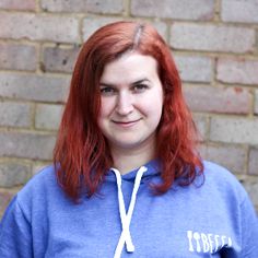 a woman with red hair wearing a blue hoodie and smiling at the camera while standing in front of a brick wall