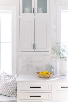 a bowl of lemons sits on the counter in this white kitchen with glass cabinets