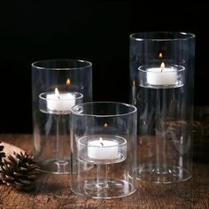 three clear glass vases with lit candles in them on a wooden table next to a pine cone