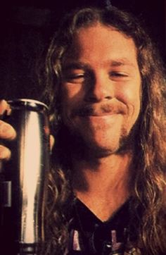 a man with long hair holding a metal cup in his right hand and smiling at the camera