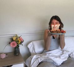 a woman holding a cake with lit candles on it in front of her face while sitting on a bed