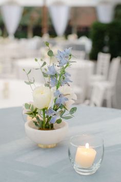 a white rose and blue flowers are in a bowl on a table with a lit candle