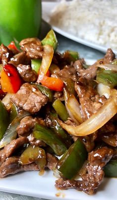 a white plate topped with beef and peppers next to rice on a tablecloth covered table