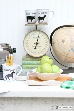 a bowl of green apples sitting on top of a table next to books and a clock