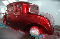 an old red car is covered with plastic wrap and some decorations on the floor in front of it