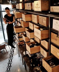 a woman standing in front of a wall full of wine bottles and wooden crates with labels on them