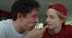 two men are eating food together in an office setting, one is wearing a red baseball cap and the other wears a white t - shirt