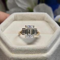 an engagement ring with three emerald cut diamonds on it in a white box next to flowers