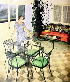 a woman standing in the middle of a living room with green chairs and a table