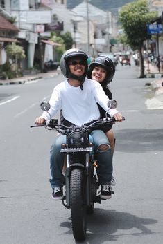 a man and woman riding on the back of a motorcycle down a street with buildings in the background