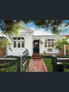 a white house with a black gate and brick walkway leading to the front door is surrounded by greenery