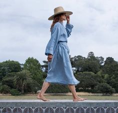 a woman in a blue dress and hat walks along a wall while talking on her cell phone