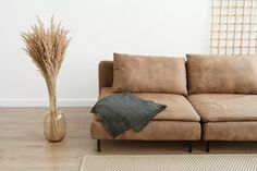 a brown couch sitting on top of a wooden floor next to a vase filled with dry grass