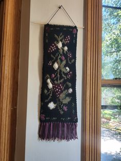 a black wall hanging with flowers and tassels on the side by a window