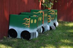 three plastic train cars sitting in the grass near a red wall and tree trunk with yellow flames painted on it