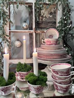 a table topped with plates and bowls filled with green plants next to two lit candles