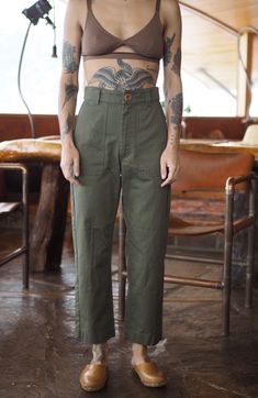 a woman with tattoos standing in front of a wooden table and wearing khaki pants