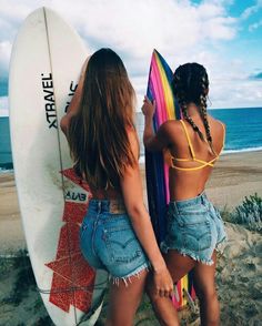 two women standing next to each other holding surfboards on the beach with ocean in background