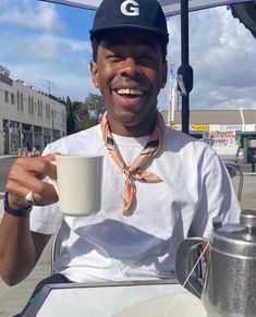 a man sitting at an outdoor table holding a coffee cup and smiling for the camera