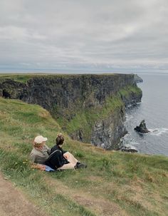 two people are sitting on the edge of a cliff overlooking the ocean and cliffs below