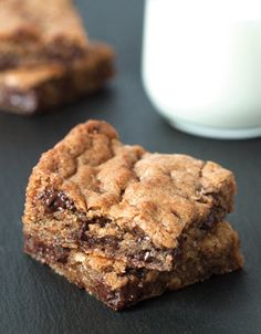 two chocolate chip cookies next to a glass of milk on a black surface with one cookie in the foreground