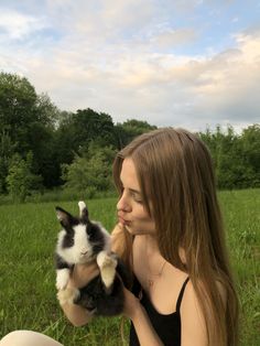 a woman is holding a small rabbit in her hands while sitting on the grass with trees in the background