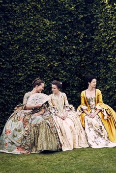 three women in period dresses sitting on the grass near a wall with ivy growing behind them