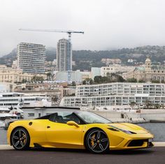 a yellow sports car is parked in front of the water and some buildings with a crane in the background