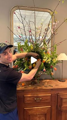 a man standing in front of a wooden dresser holding a vase with flowers on it