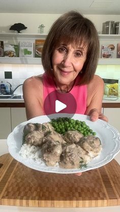 a woman holding a plate of food with meatballs and peas on it, in front of a kitchen counter