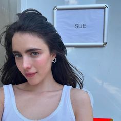 a woman with long dark hair wearing a white tank top and silver ear studs