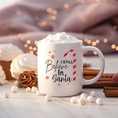 a white coffee mug with the words i still believe in santa on it next to cinnamon sticks and cupcakes