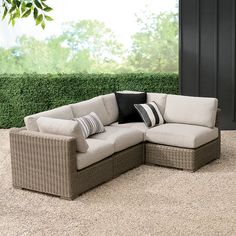 an outdoor sectional sofa with pillows on it in front of a green wall and shrubbery