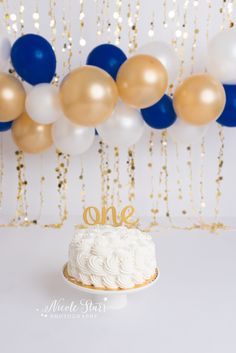 a white cake sitting on top of a table next to balloons and a string of gold and blue streamers