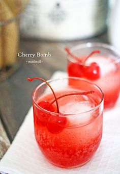 two glasses filled with red liquid and cherries on top of a white table cloth