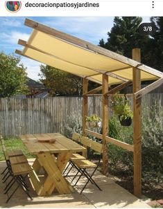 a wooden table and chairs under an awning next to a fence with trees in the background