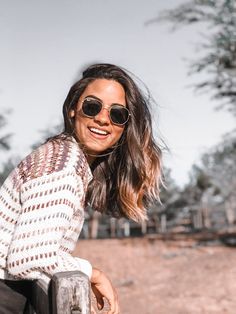 a woman wearing sunglasses leaning on a rail in front of a dirt field and trees