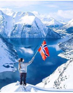 a person holding a flag on top of a snow covered mountain overlooking a lake and mountains