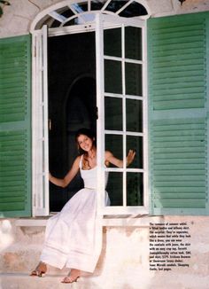 a woman in a white dress is standing by a window with green shutters and smiling