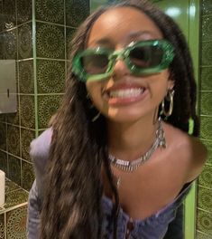 a woman wearing sunglasses in front of a green tiled wall with the reflection of her face