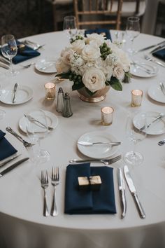 a white table with blue napkins, silverware and flowers in a gold vase