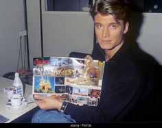 a man sitting at a desk holding up a magazine with pictures on it and looking into the camera