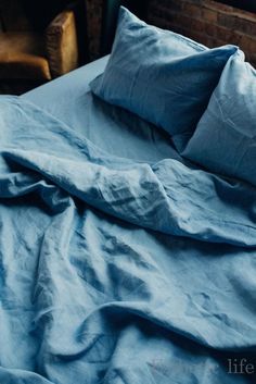 an unmade bed with blue sheets and pillows on top of it, in front of a brick wall