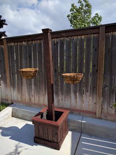 an outdoor fountain with two hanging baskets on the top, and a wooden fence behind it