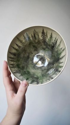 a hand holding a white bowl with green plants in the center and trees on it