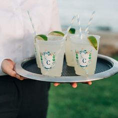 two glasses on a tray with straws and limes in them, one being held by the other