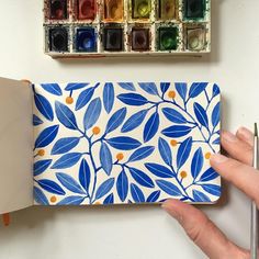 a hand holding a notebook with blue leaves on it and watercolors next to it