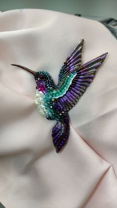 a purple and green hummingbird brooch sitting on top of a white cloth covered pillow